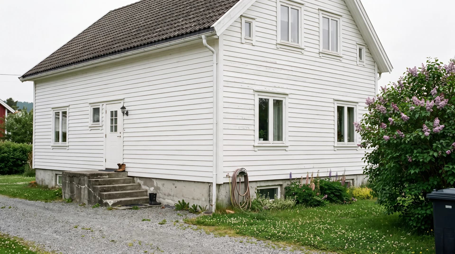 Nymalt hvit enebolig i tre en overskyet sommerdag, med grusoppkjørsel, hage og syrin i blomst.